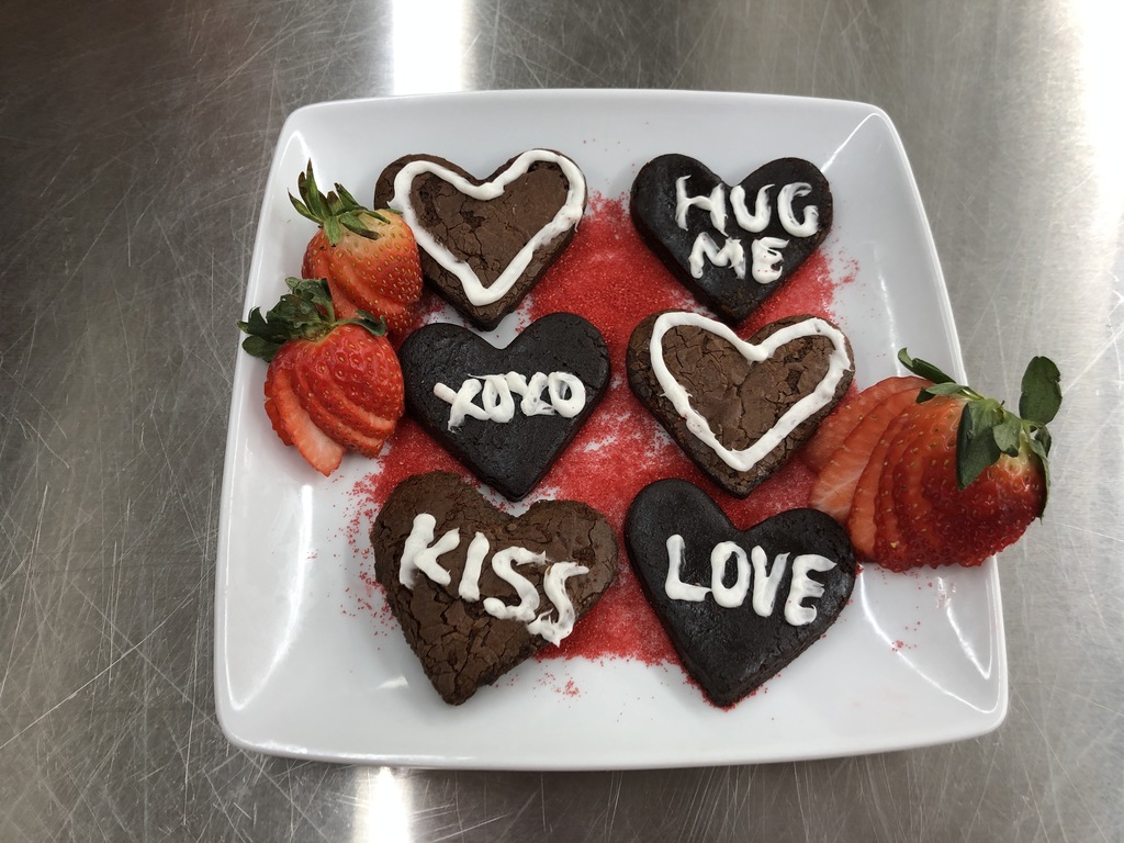 Advanced Foods class is divided up into groups and has three brownies with a description for you to vote for ONE. Please vote by "liking" the best brownie. Thank you for your help in choosing the Best Brownie! (Remember to look at all three posts before you vote.) Brownie B: Be Mine Brownies: Our Be Mine Brownies are six individual, mini heart-shaped brownies. Each brownie has a form of frosting decor, some piped with a heart, others topped with cute sayings like "XOXO". They are laying on the red sugar sprinkles along with fanned strawberries on the edges. (Chocolate Brownie, Strawberries, Sprinkles, White Whipped Frosting)